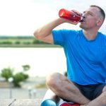 A man in a blue shirt sitting outdoors on an exercise mat, drinking from a water bottle.