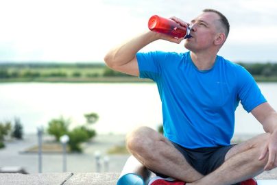 A man in a blue shirt sitting outdoors on an exercise mat, drinking from a water bottle.